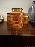 Three vintage Hornsea Saffron ceramic kitchen jars with dark wood lids placed on a wooden surface. The jars show a circular retro pattern in orange and brown colors.