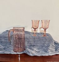 Pitcher and two goblets displayed on blue cloth, showing the swirled texture and pink hue of the glassware