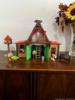 Front view of vintage Playmobil barn set with red roof, green doors, two farmer figures, pig, cow, wheelbarrow, birdhouse with birds, and a small side building.