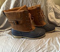 Side angled view of pair of brown and black women's winter boots with lace-up front and ankle strap, placed on beige fabric surface.