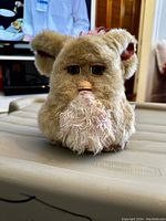 Front view of 2005 Furby Emoto Tronic showing sand-colored plush fur, pink belly, green eyes, pink tuft on head, and beak damage.