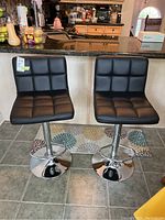 Front view of two matching black upholstered bar stools with tufted seat and back cushions and chrome metal pedestal bases with footrests.