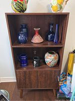 Front view of the brown laminate bookshelf with decorative ceramic and glass items on the shelves and top. Shows open shelf with assorted vases and a closed cupboard below.