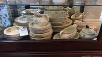 Overall assortment of Crown Ducal Florentine china pieces stacked and grouped on a glass shelf showing plates, gravy boat, saucers, cups, bowls.