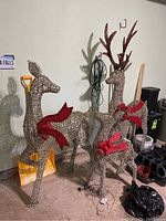 Three deer lawn figures with LED lighting visible in garage setting, each with red bows, made of woven material, shown with shovels and other garage items