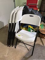 Four white plastic folding chairs with black metal frames stacked and arranged in a garage setting, showing dirt and wear on seats and backs.