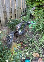 Two steel bird garden art figures standing among garden plants and a wooden fence background. The birds have detailed metal wings and necks with painted black and bronze finish.