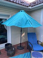 Half patio umbrella open, showing blue canopy, black metal ribs, and round stand base, set on outdoor deck near blue cushioned chair.