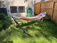 Wide shot of striped hammock mounted on black metal frame in grassy backyard next to a wooden fence and garden hose.