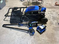 Lawn mower, string trimmer, battery, and charger arranged on garage floor showing overall condition and included items.