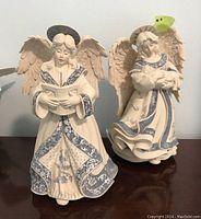 Two musical composite angel figurines shown together on a wooden surface against a wall, featuring detailed wings and robes with blue and white patterns.