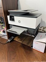 White and gray HP OfficeJet Pro 9015 inkjet printer placed on a metal and composite stand with a wood-like platform. Printer shows control panel and paper tray.