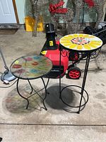 Two steel side tables on concrete floor, one with glass top featuring multi-colored leaf pattern, other with sunflower mosaic tile top and black curved metal base.