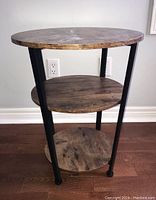 Front and side angle photo showing three circular wooden shelves supported by black metal tubular legs on a hardwood floor.