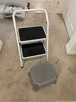 Metal folding two-step stool with black anti-slip steps and gray plastic step stool on carpeted floor.