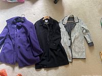 Three jackets laid flat on carpet: purple BASLER wool jacket, black vintage Novelti Laura jacket, reversible white and black Spanner jacket