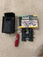 Overview of Bushnell binoculars with box, soft case, and Swiss Army pocket tool on carpet.