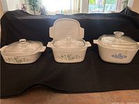 Three ceramic baking dishes shown side by side on black cloth backdrop with glass lids. Two dishes have green floral patterns, one dish has blue floral pattern.