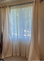 Window with sheer white curtains and cream-coloured curtains hanging on metal grommets from a curtain rod, visible through window light.