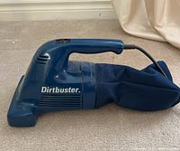 Side view of blue Black & Decker Dirtbuster corded handheld vacuum with attached dust collection bag, on carpeted floor.
