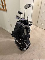 Wilson branded black and gray golf bag with rain cover partially attached, standing upright filled with golf clubs, showing front and side views of bag compartments and club heads.