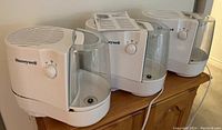 Three white Honeywell humidifiers on a wooden surface, showing side and front with control dials and clear water tanks, along with a manual placed on top.