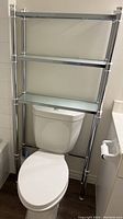Metal shelving unit positioned over a toilet, with three glass shelves attached to a silver metal frame.