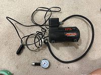 Mini-mite air compressor with attached hose and power cord with car adapter plug, and a separate air pressure gauge on beige carpet.
