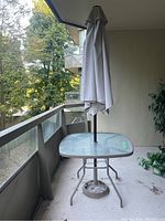 Front view of outdoor square glass table with beige umbrella mounted in center. Shows table top and umbrella closed, located on balcony floor.