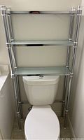 Full view of silver metal shelving unit with three glass shelves installed over a toilet in a bathroom.