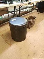 Rectangular woven basket with handles and round dark brown storage bin with vinyl top shown on wooden floor near folding table.