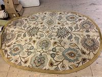 Full view of the oval wool carpet showing the entire floral pattern and mustard yellow border on a wooden floor with some boxes partially visible nearby.