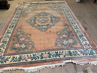 Full view of rectangular wool carpet with central floral medallion and surrounding floral border on peach background, fringed edges visible.