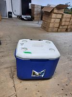 Front view of blue and white Coleman cooler with visible torn sticker and broken handle.