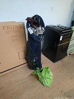 Full view of blue golf bag standing upright, partially filled with golf clubs, some wrapped in protective sheets, with a green reusable shopping bag on the floor beside it.