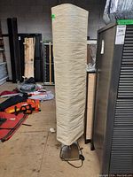 Full front view of a tall rectangular floor lamp with beige paper shade on a metal base in a warehouse environment.