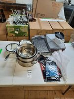 Wide view showing various kitchen pots, pans, metal trays, clear pitcher with blue lid, kitchen food tent, folded plastic tablecloth, paper tablecloths, and assorted kitchen utensils in boxes.