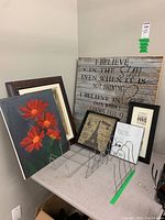 Photo showing wood panel with inspirational quote, floral painting on canvas with red flowers, and several framed prints leaning against the wall on a table.