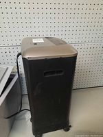 Side and front view of tall black and silver paper shredder with wheels, power cord visible, placed against pegboard background.