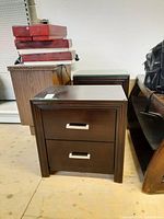 Two dark wood side tables each with two drawers, metal handles visible. Scratches and wear visible on surfaces. Located in storage area with various other furniture items around.