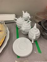 Four white ceramic tea for one sets, including teapots and saucers, along with stacked cups and saucers visible in second image.