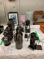 Seven metal wine bottle holders displayed on a white tablecloth showing various artistic designs.