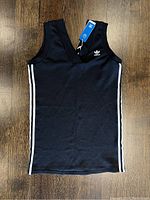 Front view of black sleeveless Adidas dress with white side stripes and trefoil logo on chest on wood floor background.