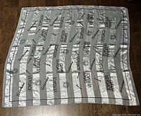 Top view of the entire scarf laid flat showing the striped silver and grey pattern with repeated brand names and decorative circular motifs.