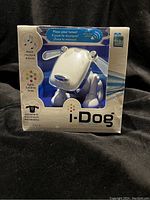Box front showing the i-Dog speaker in silver with icons indicating music play, lights, and movement