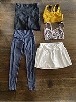 Photo showing black shorts, blue leggings, mustard yellow sports bra, floral sports bra, and white pleated skirt arranged on a wooden floor.