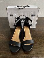 Front view of new black ankle strap heels showing toe strap and heel style with original box in background.
