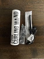 Photo showing curling wand with cylindrical white packaging tube labeled '25mm Curling Wand' on wooden surface.