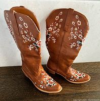 Pair of new brown leather knee-high cowboy boots with embroidered floral embellishments on shafts and toe areas shown side by side.