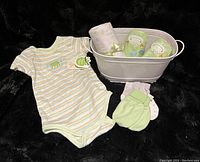 Photo showing the full lot of newborn items arranged on black background, including striped onesie, mittens, leather booties, rolled blanket, and tub.